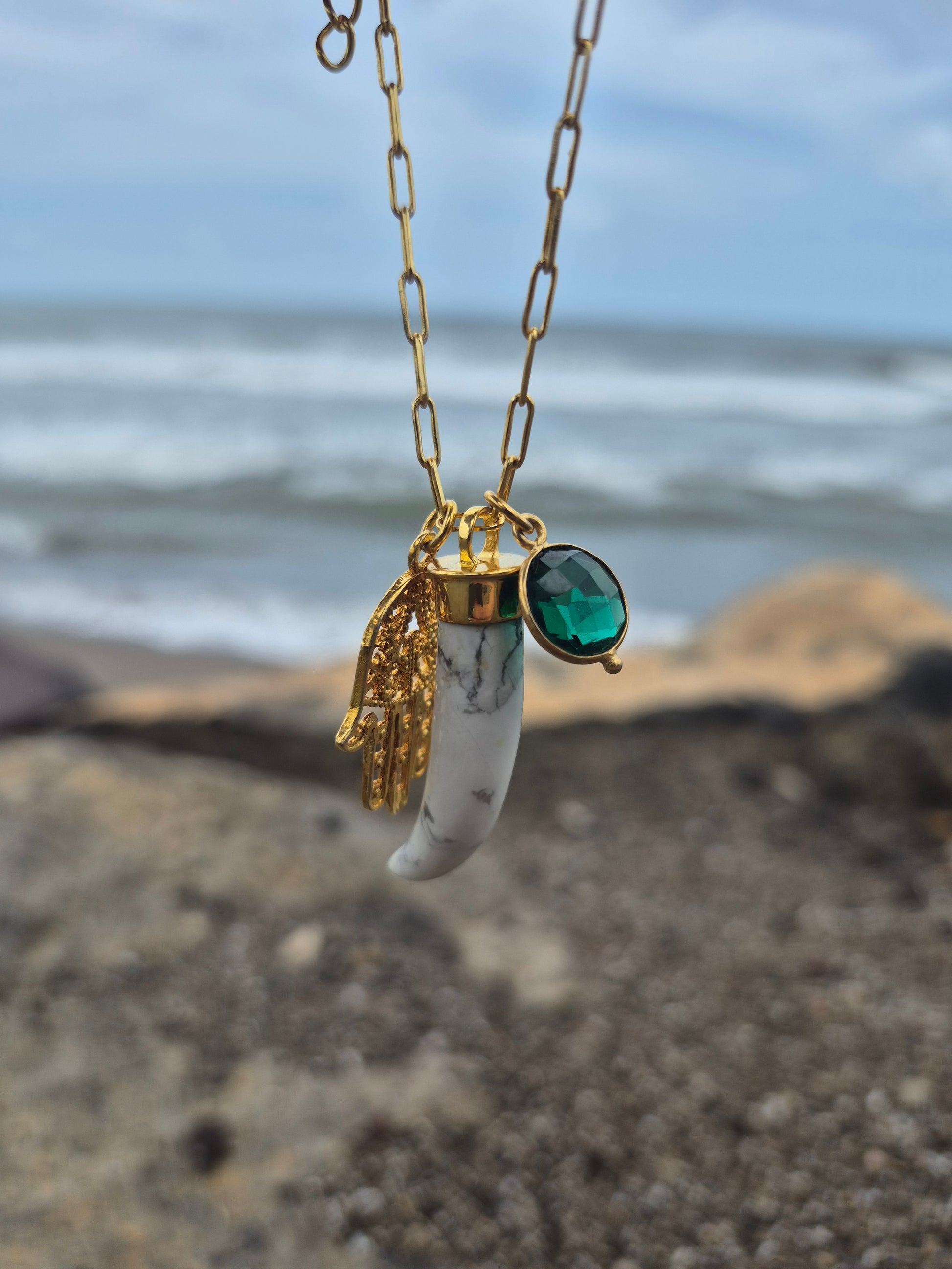 Necklace with a gold chain and pendant featuring a green gemstone and clear stone, against a blurred natural background.