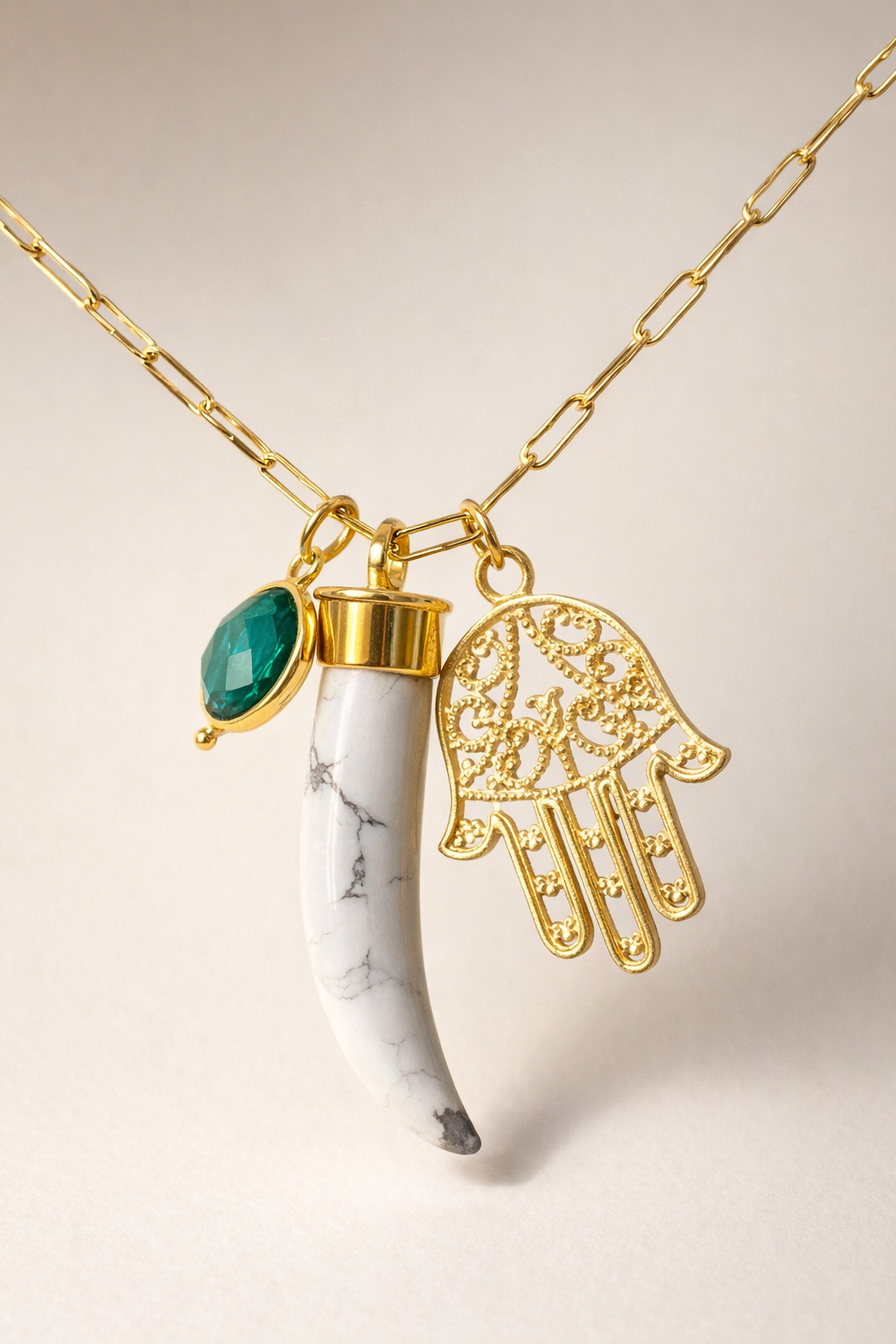 Gold necklace with a horn, green gemstone, and gold Hamsa hand pendant on a beige background